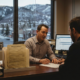 A bail bond agent works at a desk cluttered with casework and an illuminated computer screen, representing the administrative work involved in posting bail. He talks with a client on the phone while reviewing an application, with a background window revealing the high mountain desert landscape of Utah at dusk